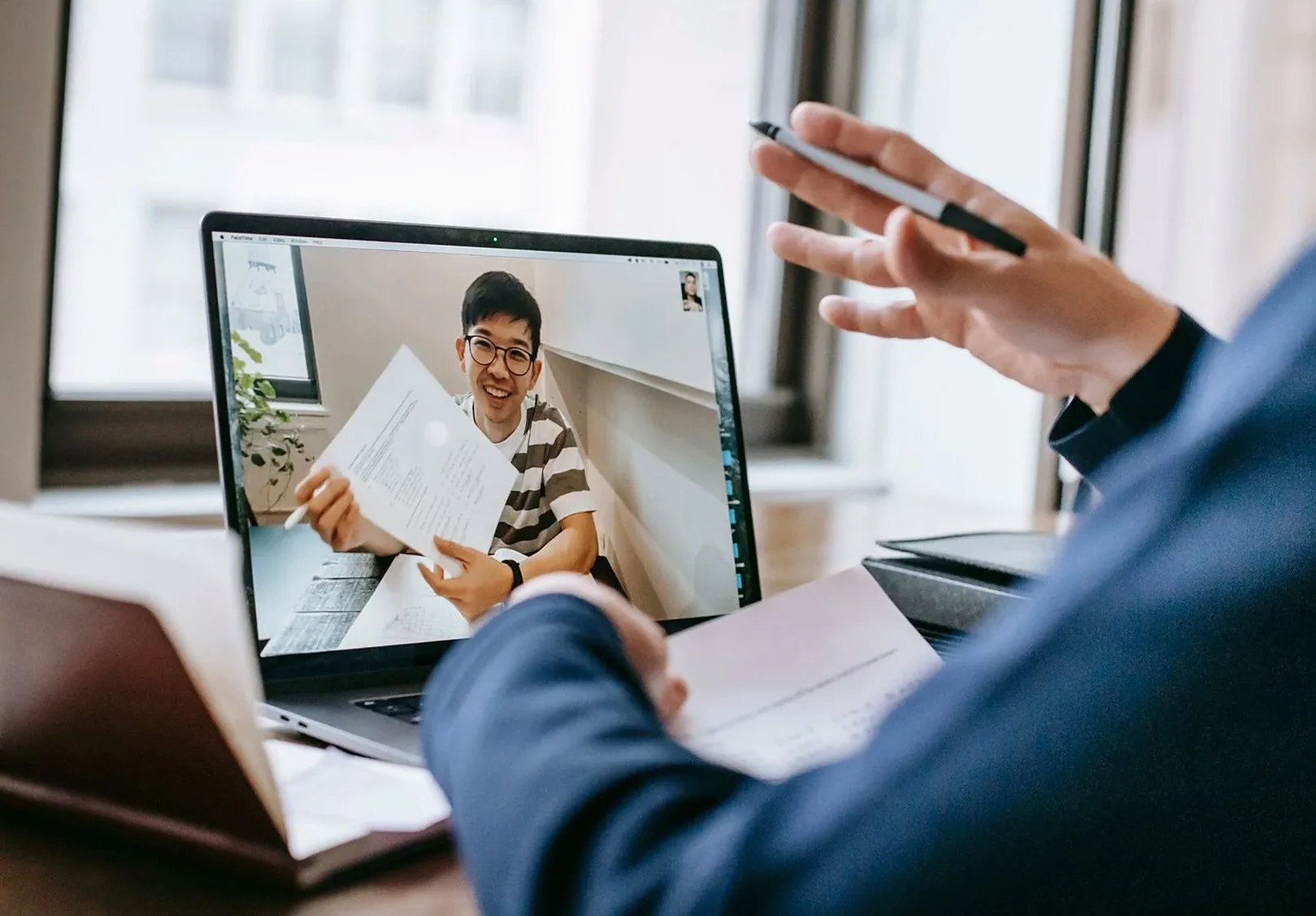 Laptop showing a video call, with a hand holding a pen in the foreground