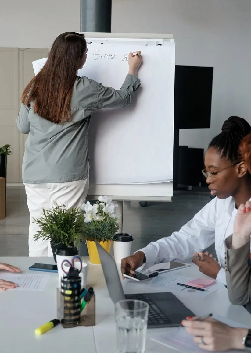 A woman writing on a flip chart while two colleagues work at a table in a meeting room
