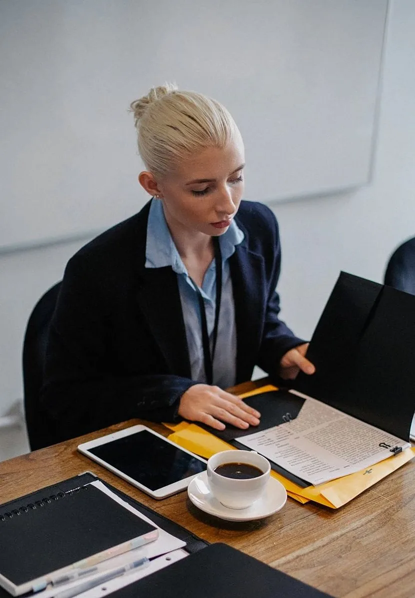 A woman in a blazer reviewing documents at a desk, with a tablet and a cup of coffee